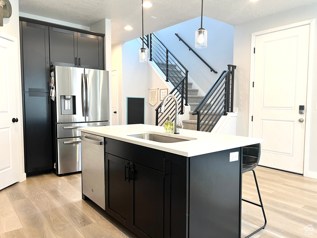 Kitchen featuring appliances with stainless steel finishes, light wood-style flooring, light countertops, a center island with sink, and pendant lighting