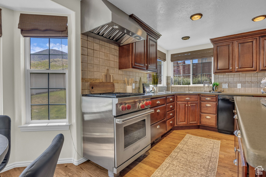 Kitchen with designer stove, wall chimney range hood, black dishwasher, tasteful backsplash, and light wood-style floors