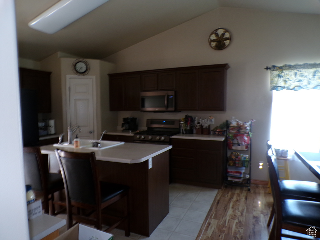 Kitchen with appliances with stainless steel finishes, lofted ceiling, light countertops, a center island with sink, and dark brown cabinets