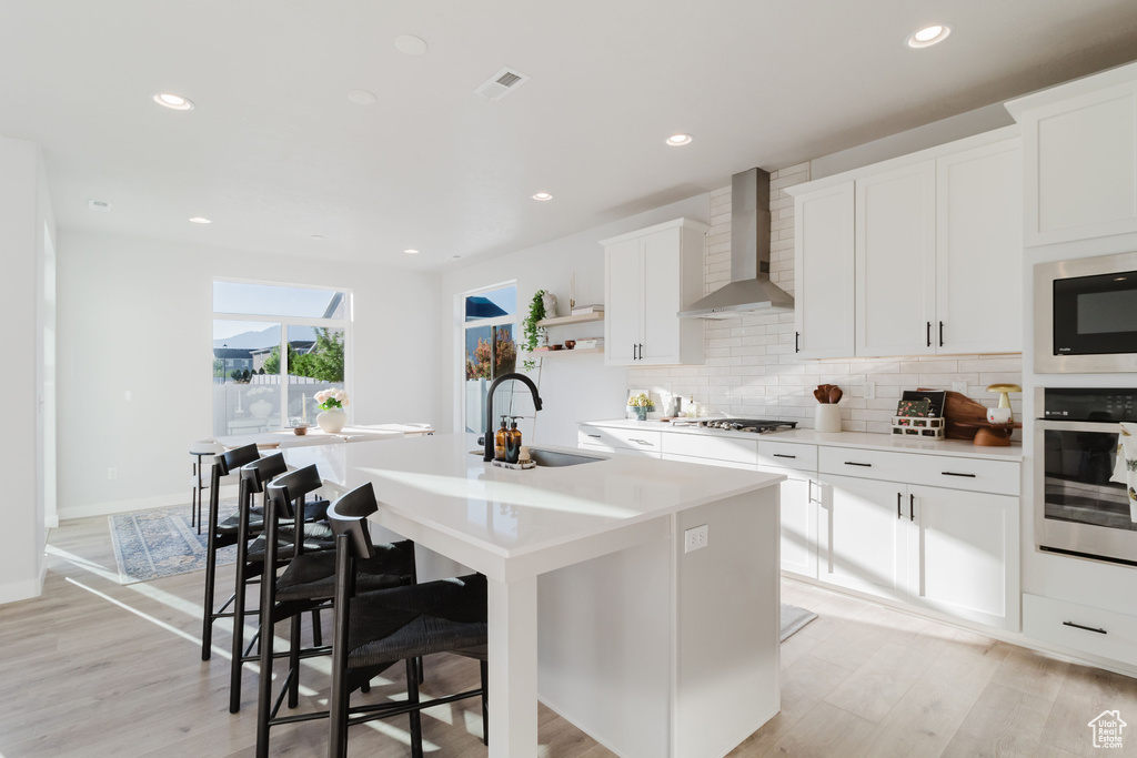 Kitchen with wall chimney exhaust hood, stainless steel appliances, a sink, a kitchen island with sink, and backsplash