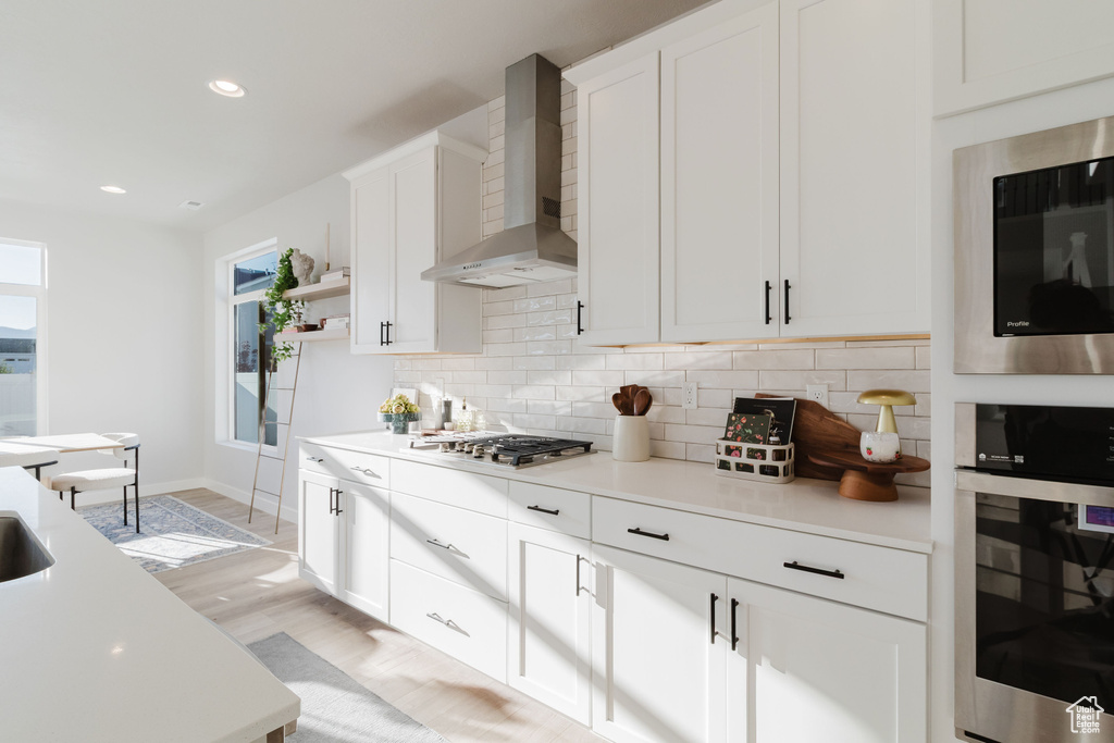 Kitchen featuring stainless steel appliances, wall chimney exhaust hood, decorative backsplash, open shelves, and light countertops