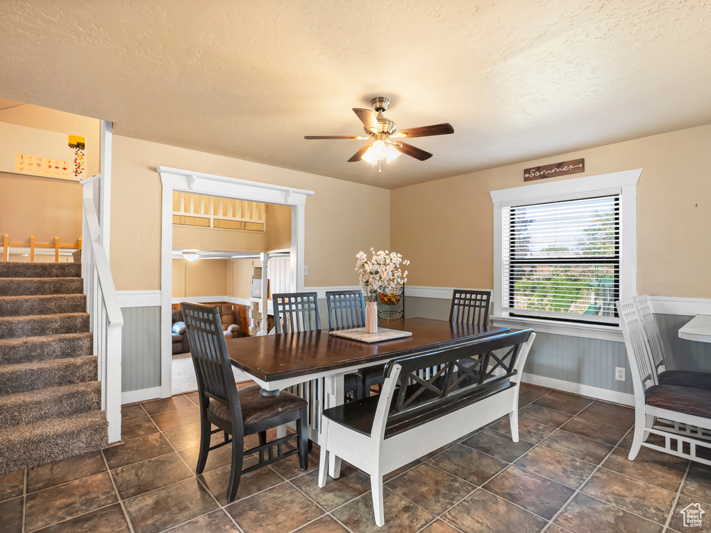 Dining room featuring a textured ceiling, wainscoting, a ceiling fan, stairs, and stone finish flooring