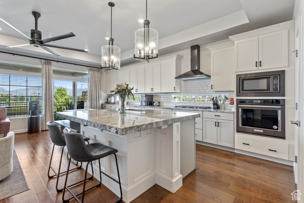 Kitchen with stainless steel appliances, wall chimney range hood, a raised ceiling, white cabinetry, and backsplash