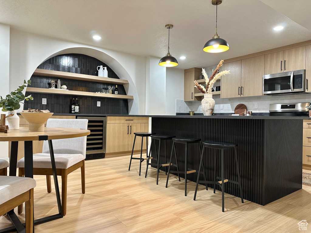 Indoor bar with stainless steel microwave, beverage cooler, decorative backsplash, light wood-style flooring, and decorative light fixtures