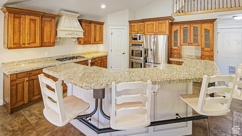 Kitchen with custom range hood, appliances with stainless steel finishes, brown cabinets, vaulted ceiling, and a breakfast bar