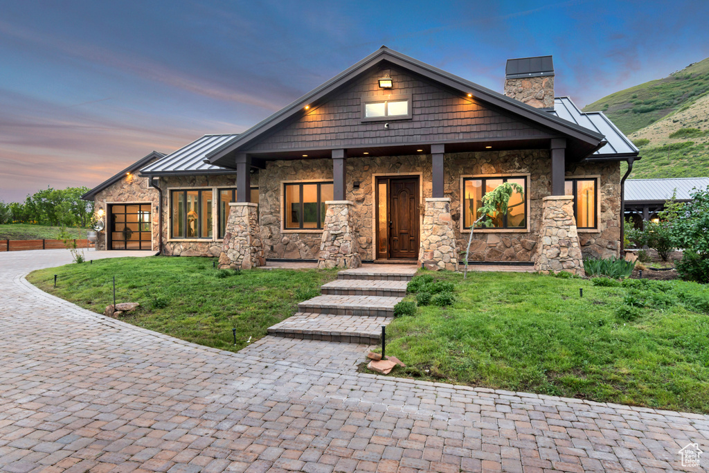 View of front facade with stone siding, a chimney, a standing seam roof, decorative driveway, and a lawn