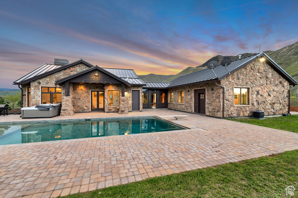Back of property with a hot tub, a standing seam roof, a patio, stone siding, and an outdoor pool