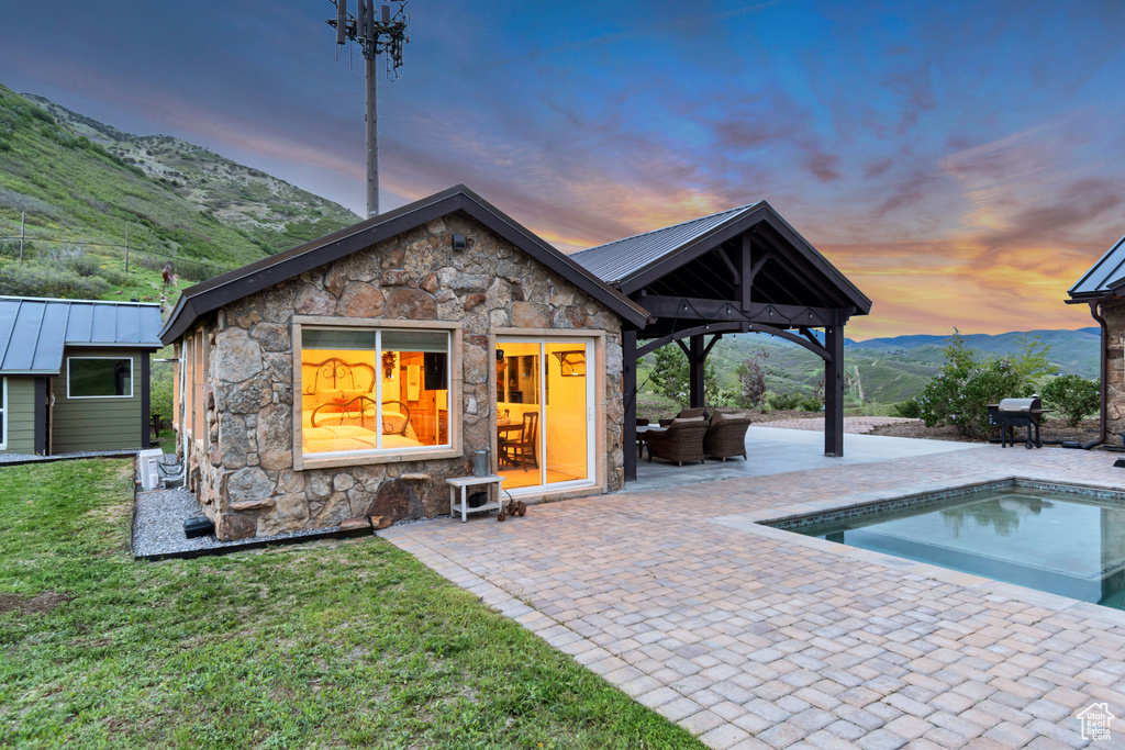 Back of house at dusk with stone siding, a mountain view, a patio area, an outdoor pool, and metal roof