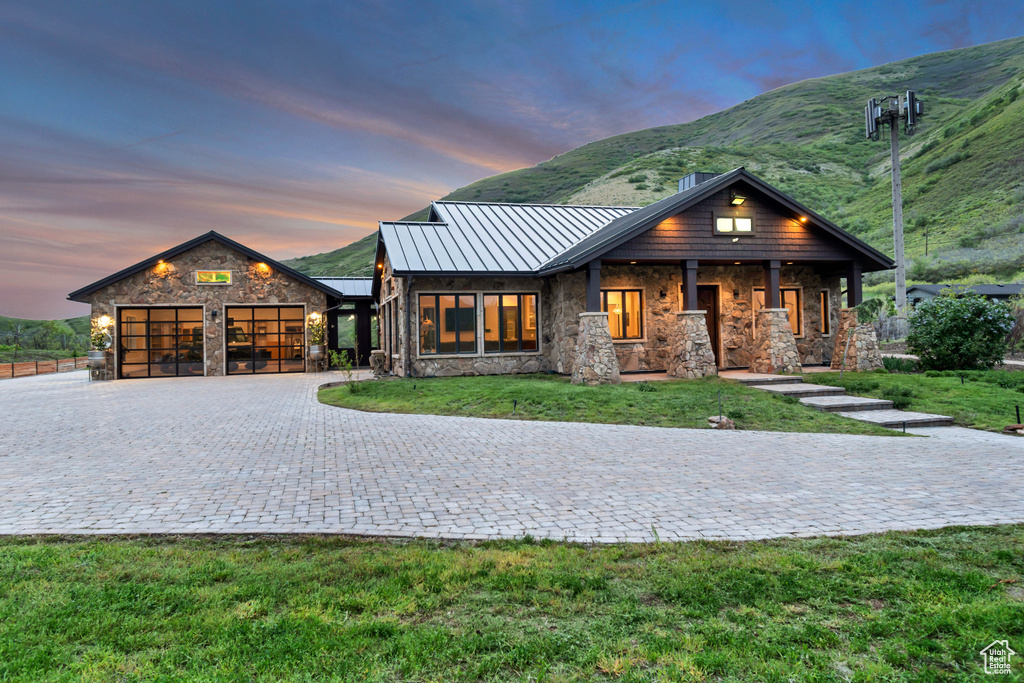 View of front facade with a standing seam roof, stone siding, decorative driveway, a garage, and a front lawn