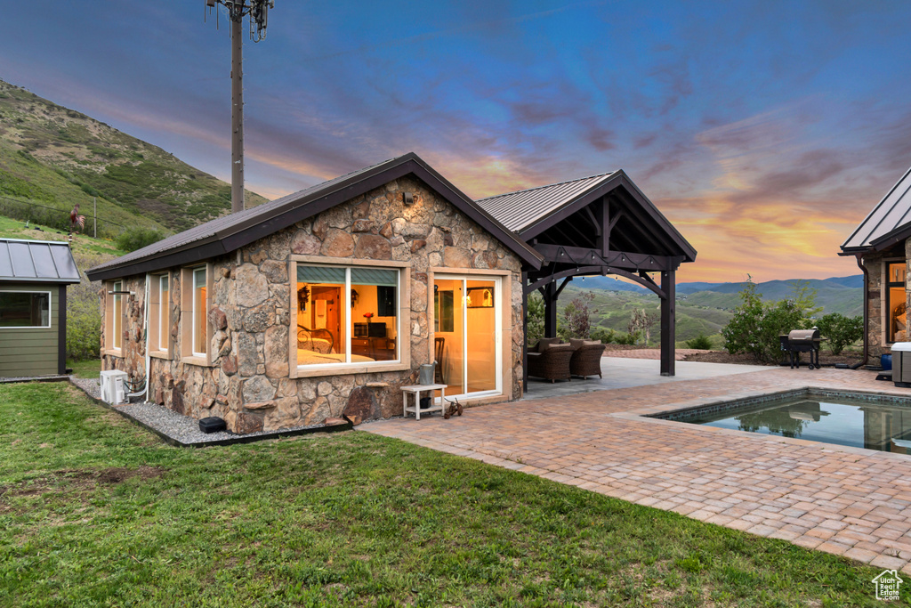 Back of house at dusk featuring stone siding, a patio, a lawn, an outdoor pool, and a standing seam roof