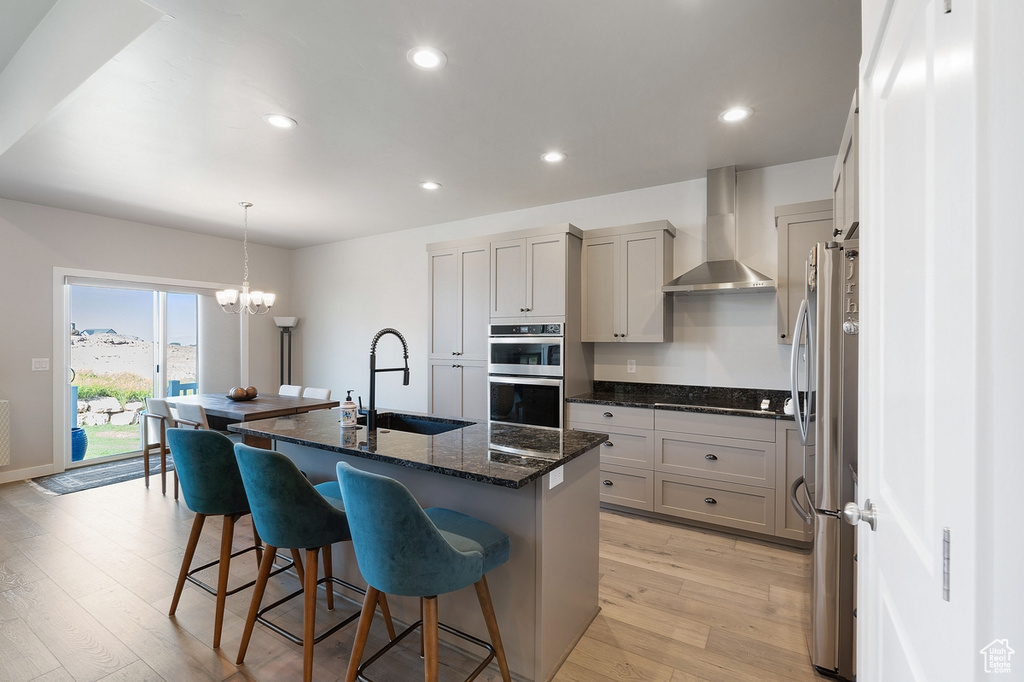 Kitchen featuring wall chimney range hood, a sink, stainless steel appliances, light wood-type flooring, and an island with sink