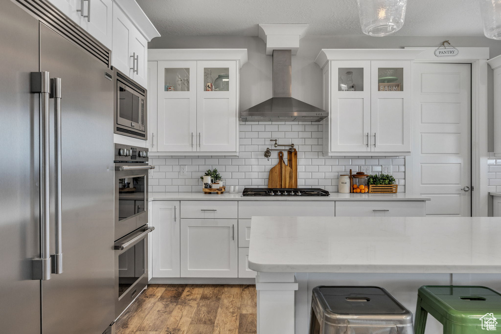 Kitchen featuring appliances with stainless steel finishes, wall chimney exhaust hood, light countertops, white cabinets, and glass insert cabinets