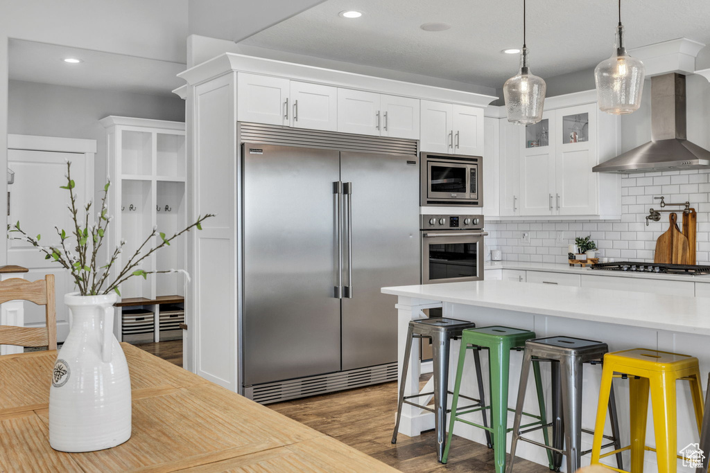 Kitchen featuring built in appliances, wall chimney range hood, light countertops, white cabinets, and recessed lighting