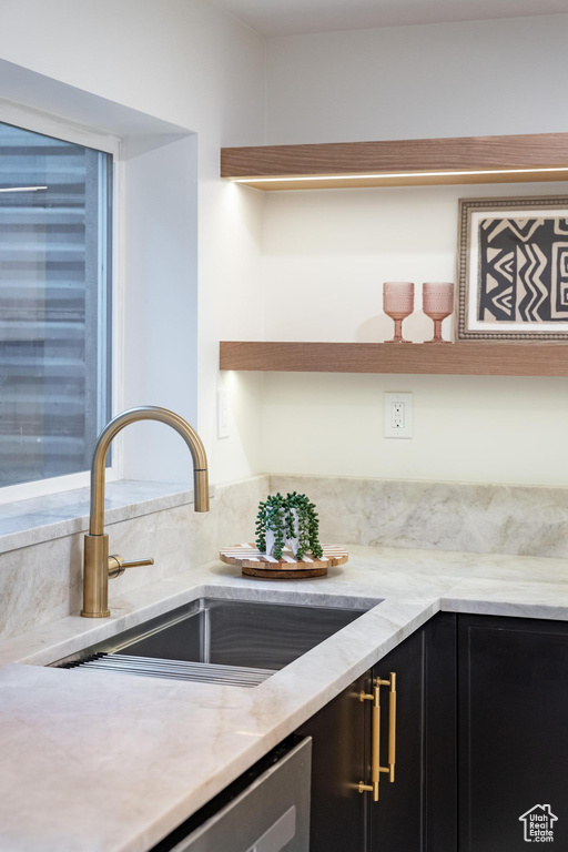 Kitchen featuring a sink, open shelves, dark cabinets, light stone counters, and stainless steel dishwasher