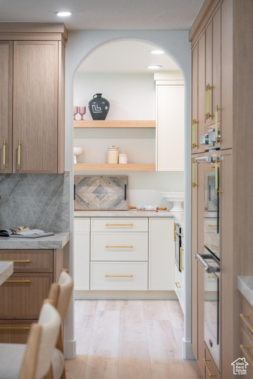 Kitchen featuring arched walkways, light countertops, light wood finished floors, stainless steel double oven, and decorative backsplash