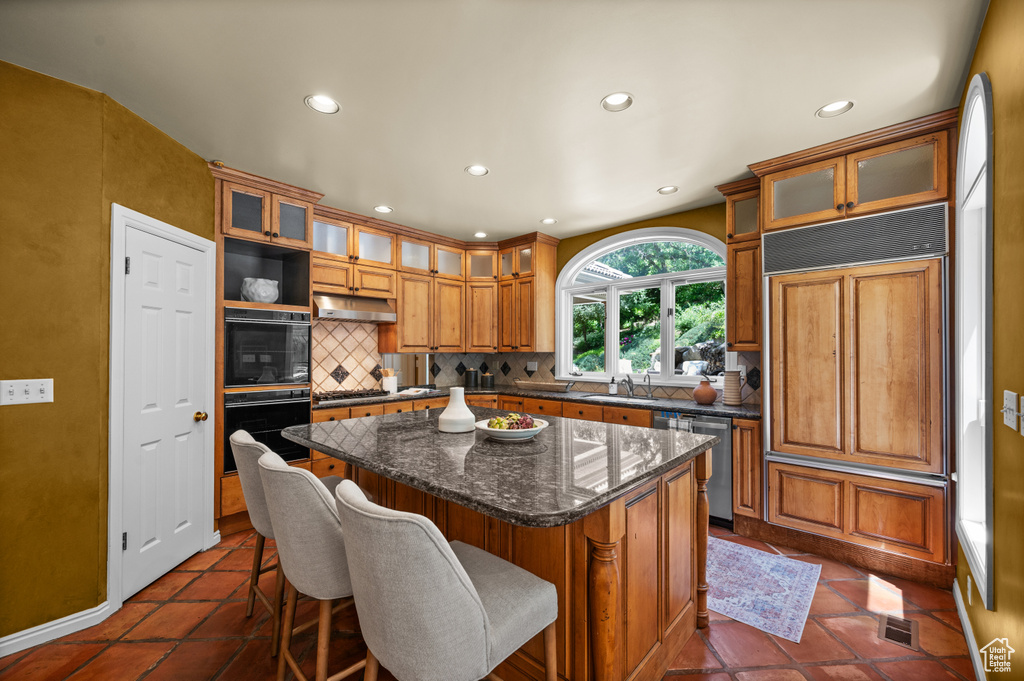 Kitchen featuring stainless steel dishwasher, under cabinet range hood, tasteful backsplash, a kitchen island, and brown cabinetry