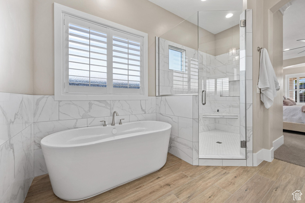 Ensuite bathroom featuring tile walls, a shower stall, wood finished floors, a soaking tub, and a wainscoted wall