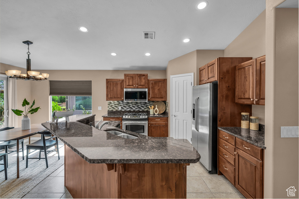 Kitchen with stainless steel appliances, backsplash, brown cabinetry, a center island with sink, and recessed lighting