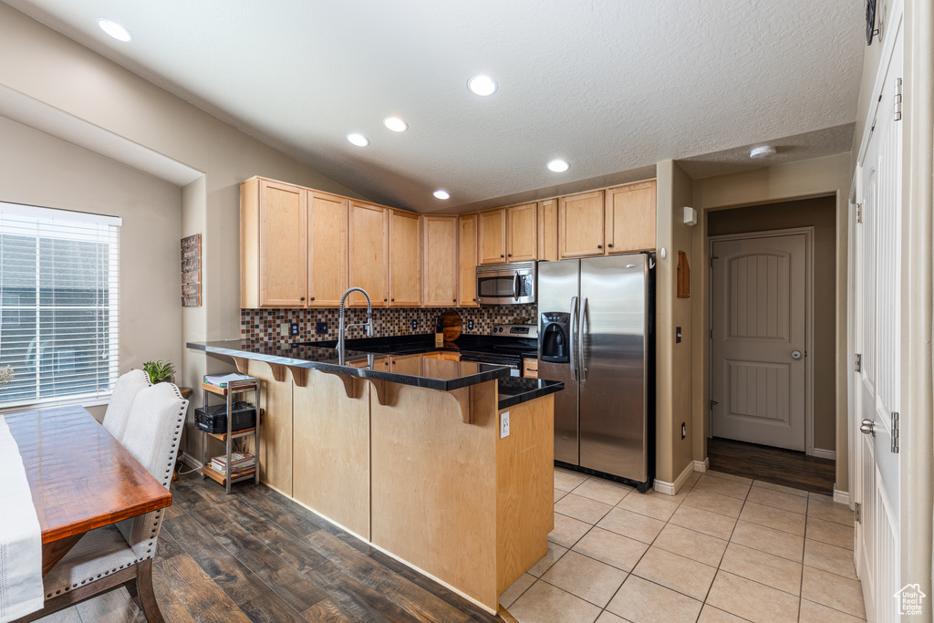 Kitchen featuring stainless steel appliances, a peninsula, light brown cabinetry, dark countertops, and recessed lighting
