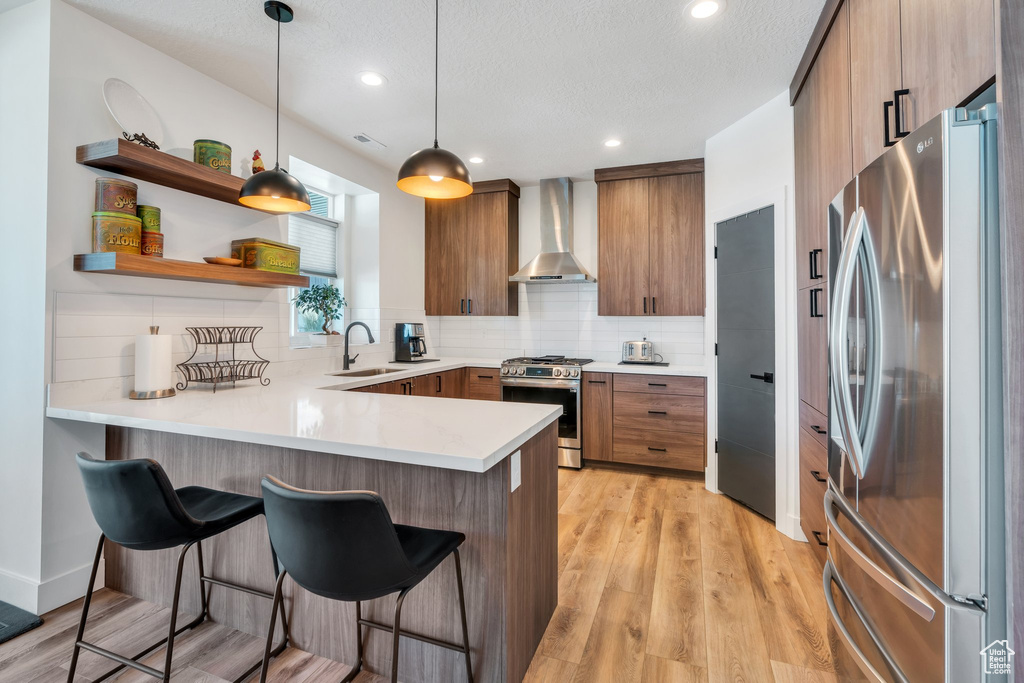 Kitchen with wall chimney exhaust hood, stainless steel appliances, a peninsula, a kitchen bar, and light countertops