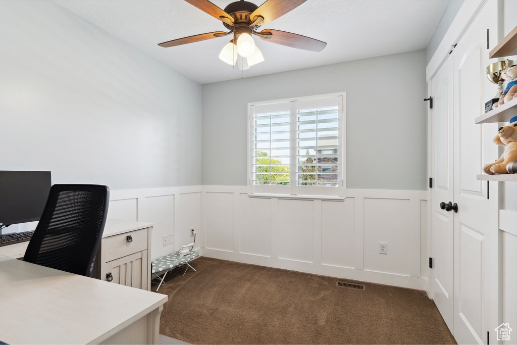 Office area with dark colored carpet, a wainscoted wall, ceiling fan, and a decorative wall