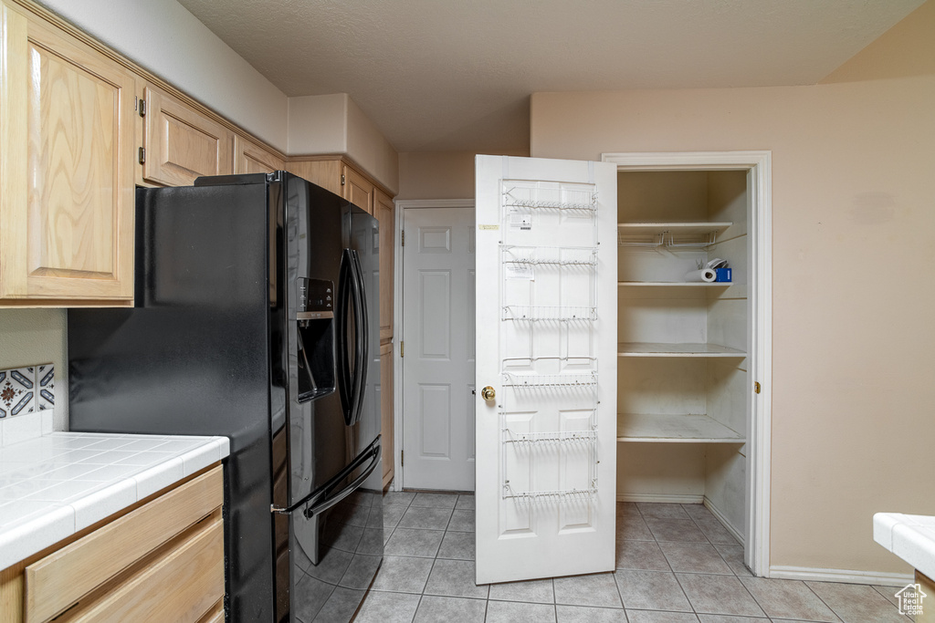 Kitchen featuring tile countertops, light brown cabinetry, black fridge with ice dispenser, light tile patterned floors, and baseboards