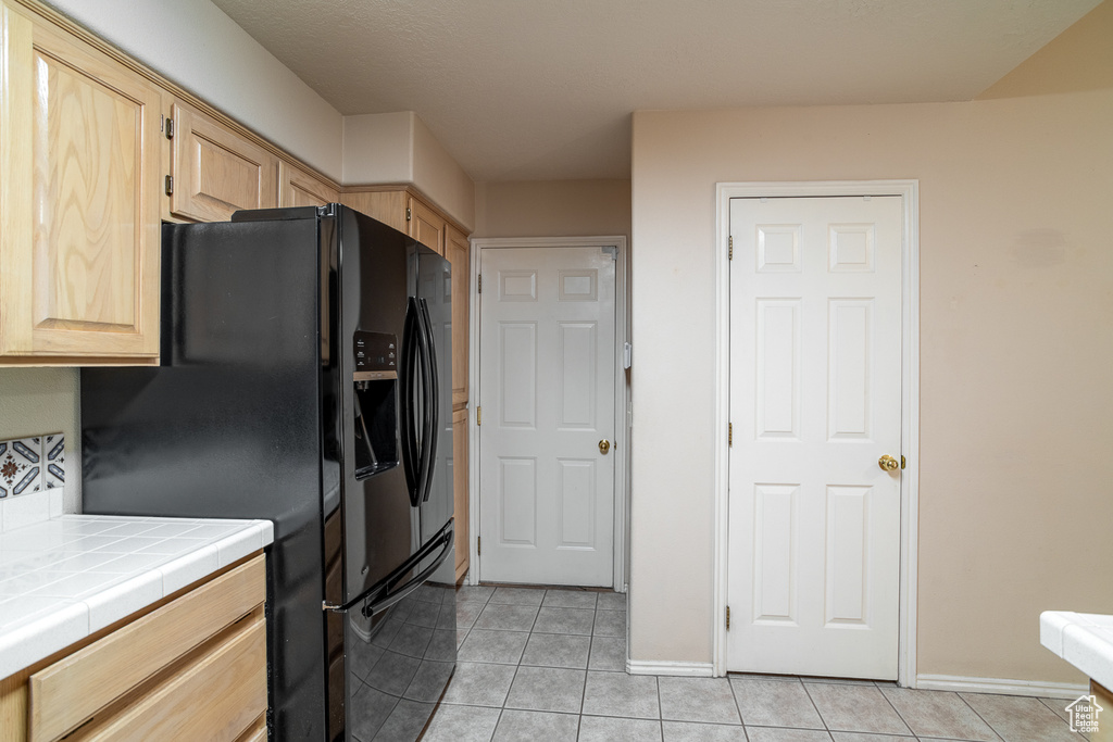 Kitchen with light brown cabinets, tile counters, black fridge with ice dispenser, light tile patterned floors, and baseboards