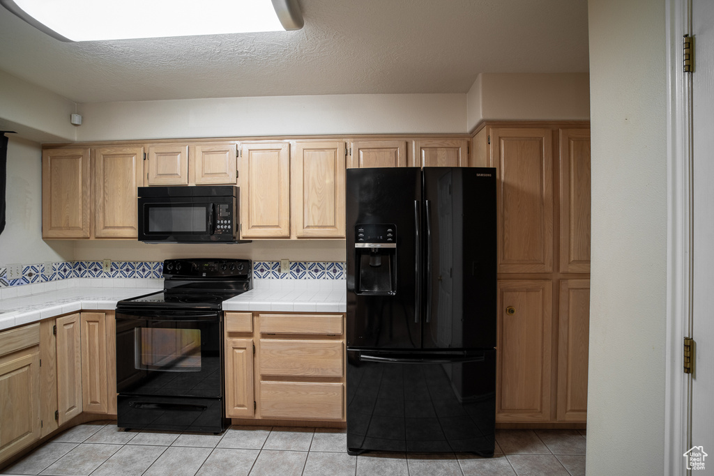 Kitchen featuring black appliances, tile countertops, light brown cabinets, and light tile patterned floors