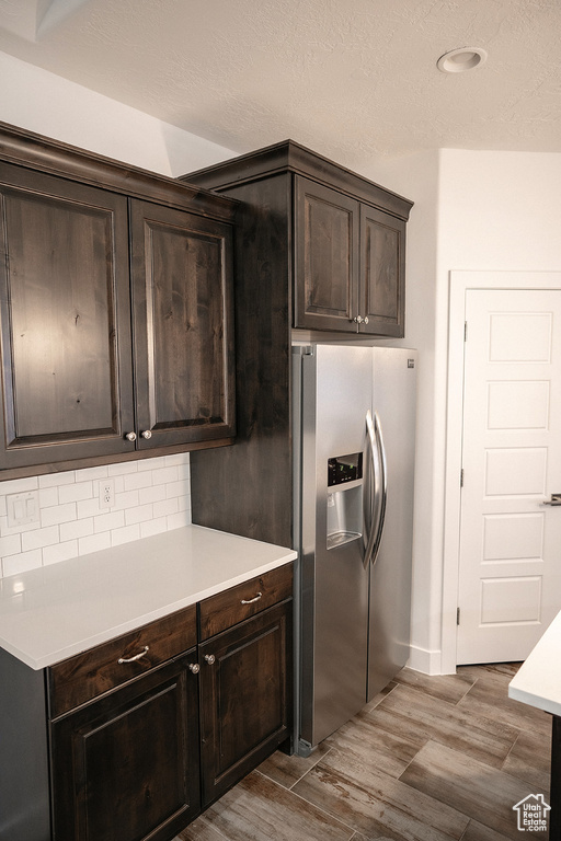Kitchen with stainless steel fridge, wood finished floors, dark brown cabinetry, light countertops, and a textured ceiling
