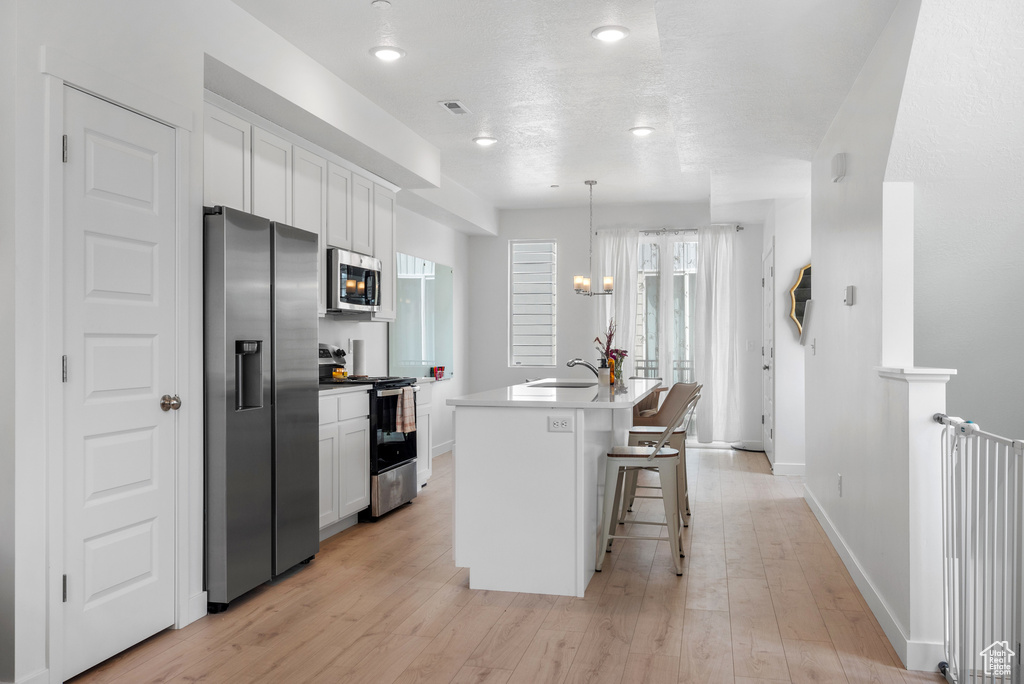 Kitchen featuring appliances with stainless steel finishes, white cabinets, an island with sink, light wood-style floors, and a kitchen breakfast bar