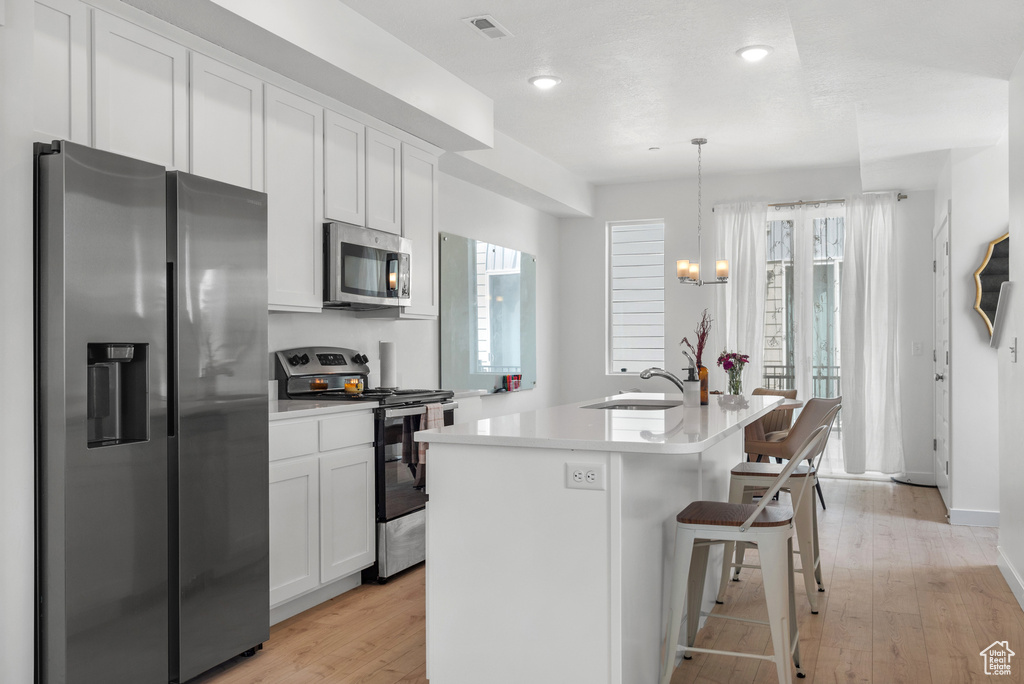 Kitchen featuring stainless steel appliances, white cabinets, an island with sink, light countertops, and light wood-style flooring