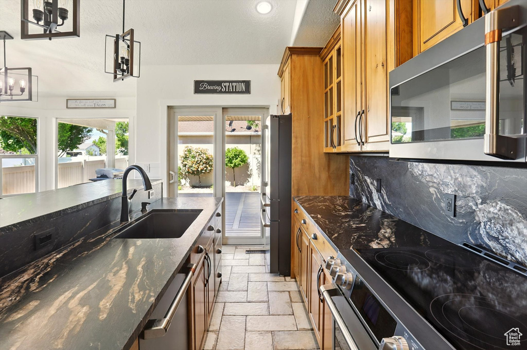 Kitchen featuring appliances with stainless steel finishes, brown cabinetry, stone tile flooring, dark stone counters, and recessed lighting