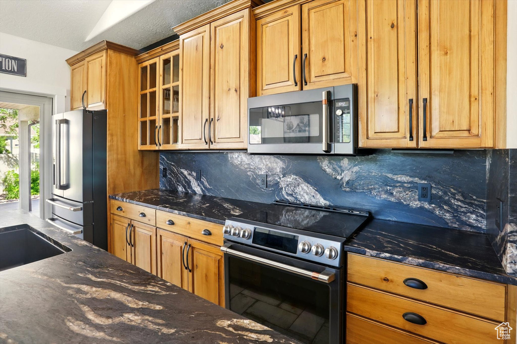 Kitchen featuring stainless steel appliances, backsplash, glass insert cabinets, dark stone countertops, and brown cabinetry
