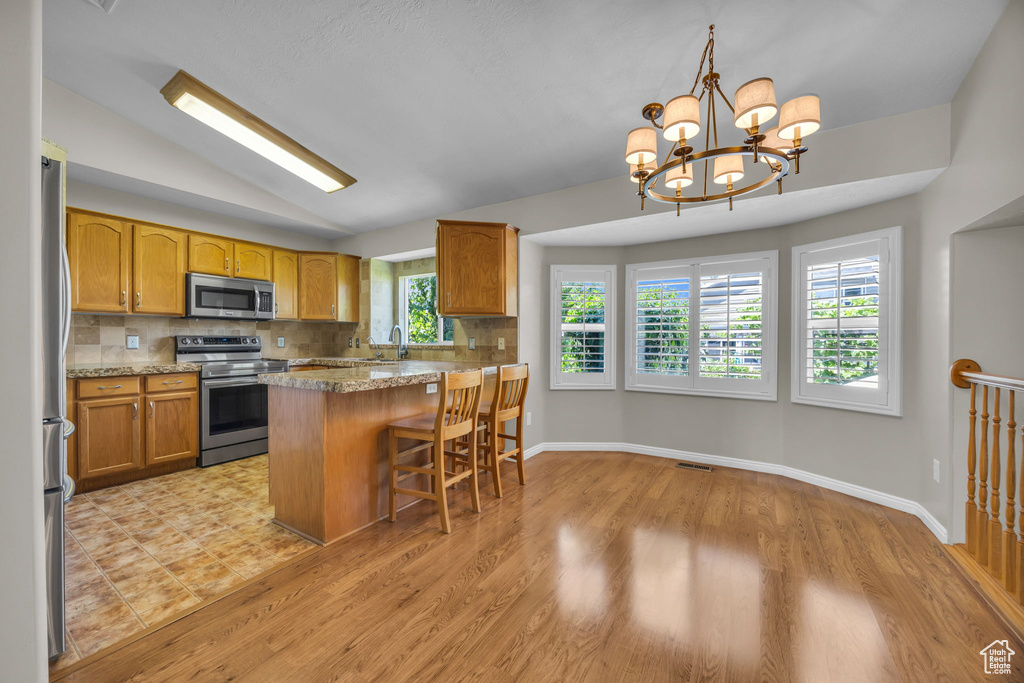 Kitchen with stainless steel appliances, backsplash, a breakfast bar, brown cabinetry, and vaulted ceiling