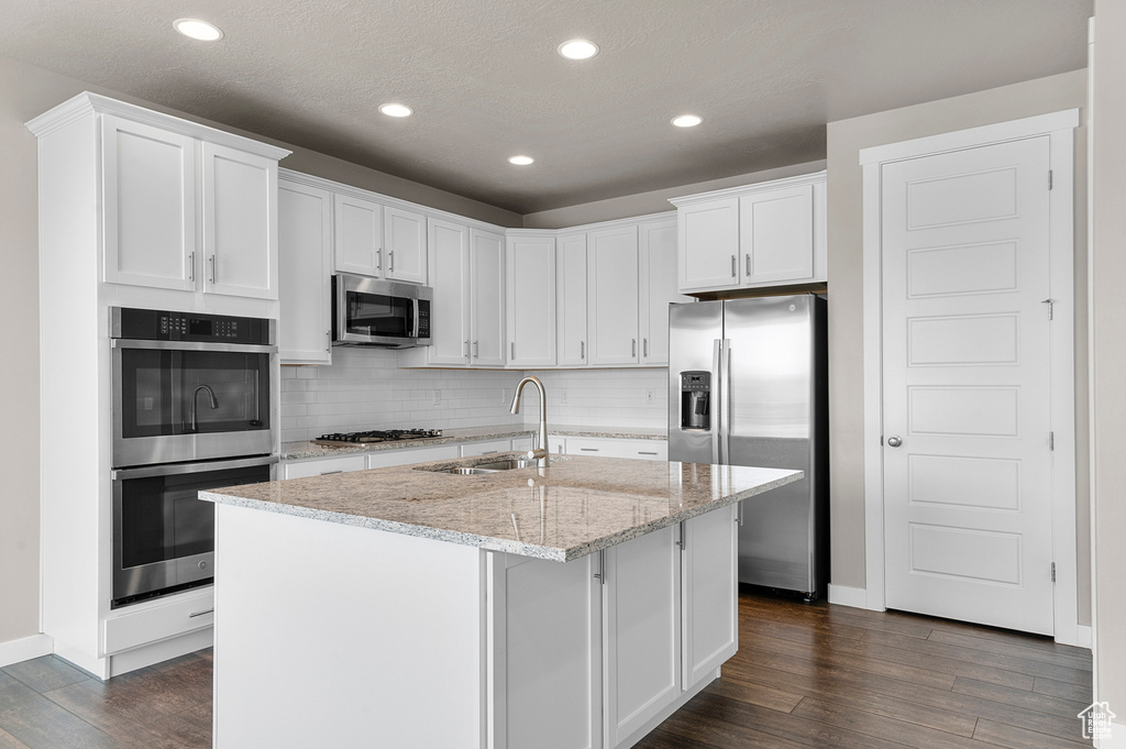 Kitchen with stainless steel appliances, dark wood finished floors, white cabinetry, a center island with sink, and backsplash
