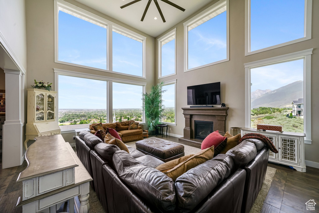 Living area featuring a towering ceiling, a fireplace with raised hearth, plenty of natural light, a ceiling fan, and dark wood-type flooring
