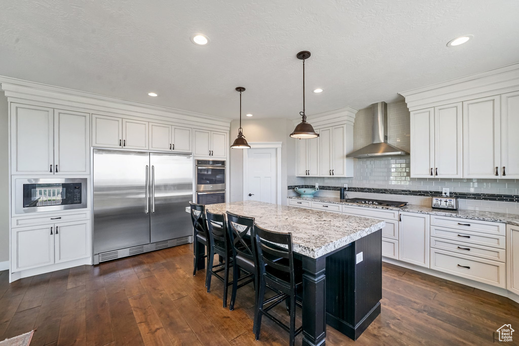 Kitchen with built in appliances, wall chimney exhaust hood, dark wood-style flooring, a kitchen bar, and tasteful backsplash