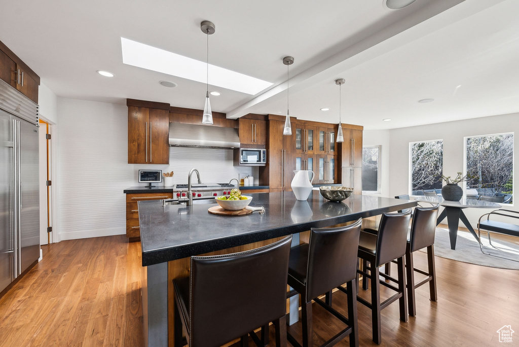 Kitchen featuring dark countertops, built in appliances, wall chimney exhaust hood, a breakfast bar, and light wood-style flooring