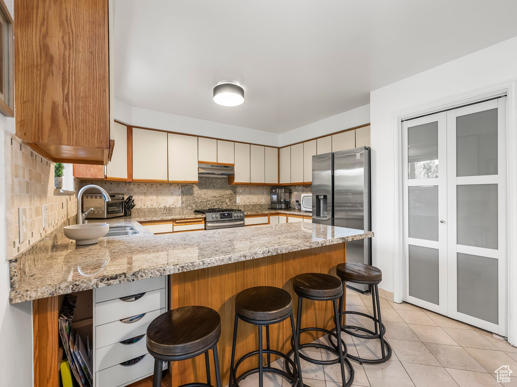 Kitchen with appliances with stainless steel finishes, under cabinet range hood, a peninsula, backsplash, and light stone counters