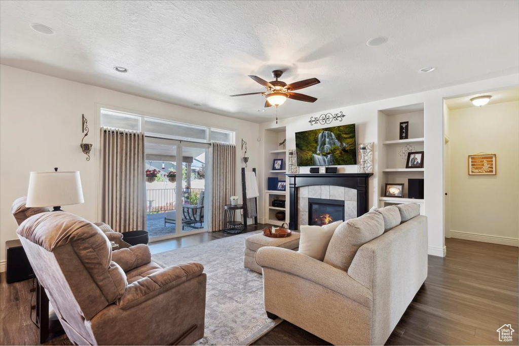Living room with a tiled fireplace, ceiling fan, dark wood-type flooring, built in shelves, and recessed lighting