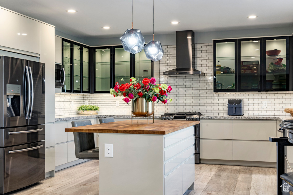 Kitchen featuring fridge with ice dispenser, butcher block countertops, wall chimney exhaust hood, gas range, and white cabinetry