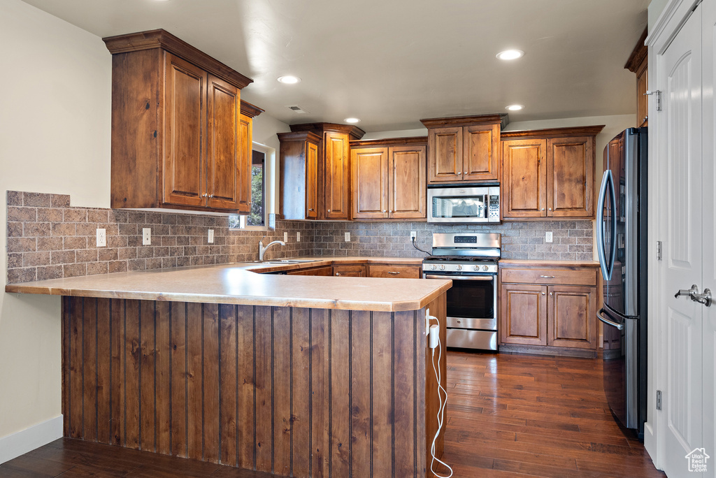Kitchen featuring appliances with stainless steel finishes, a peninsula, light countertops, brown cabinets, and dark wood-type flooring