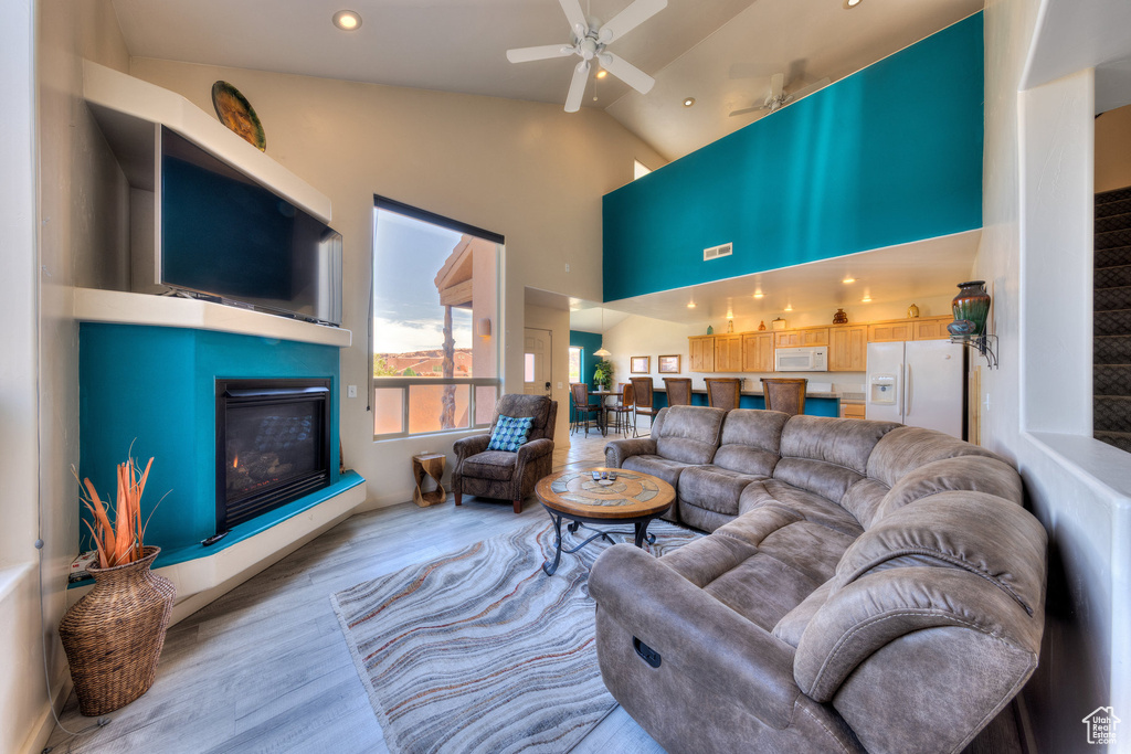 Living area with a glass covered fireplace, wood finished floors, ceiling fan, high vaulted ceiling, and recessed lighting