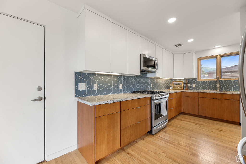 Kitchen featuring appliances with stainless steel finishes, light wood-type flooring, light stone countertops, white cabinetry, and backsplash
