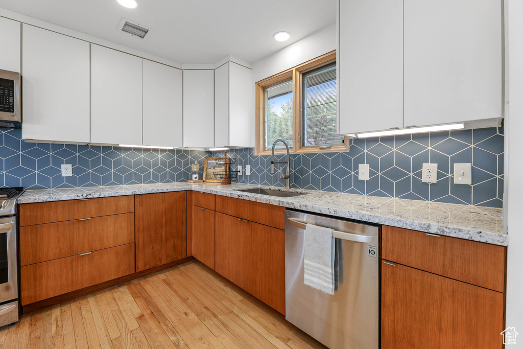 Kitchen with appliances with stainless steel finishes, light wood-type flooring, light stone counters, white cabinets, and decorative backsplash