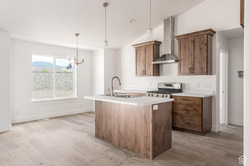 Kitchen featuring wall chimney exhaust hood, stainless steel range with gas stovetop, a center island with sink, light countertops, and light wood-type flooring