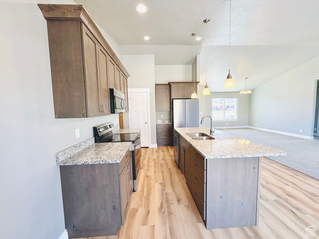 Kitchen with stainless steel appliances, light wood-style flooring, a center island with sink, hanging light fixtures, and light stone counters