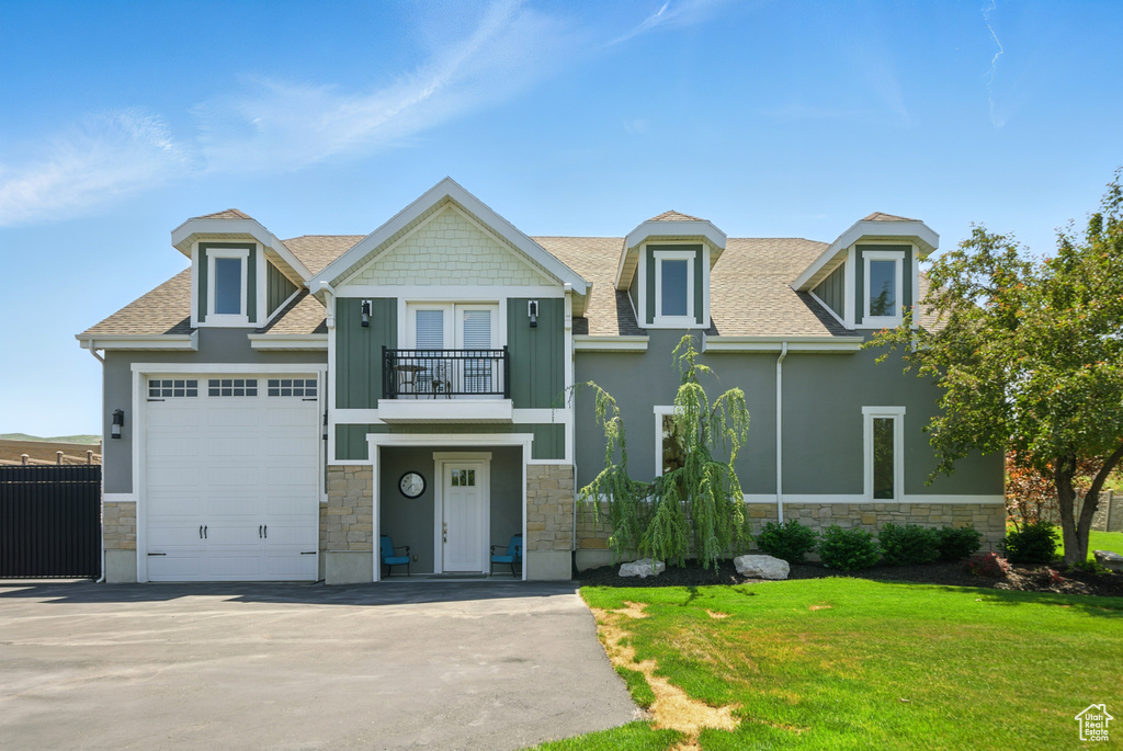 View of front of house featuring stone siding, a balcony, a shingled roof, a front lawn, and driveway
