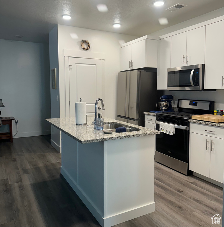 Kitchen featuring stainless steel appliances, dark wood-type flooring, light stone countertops, an island with sink, and white cabinets