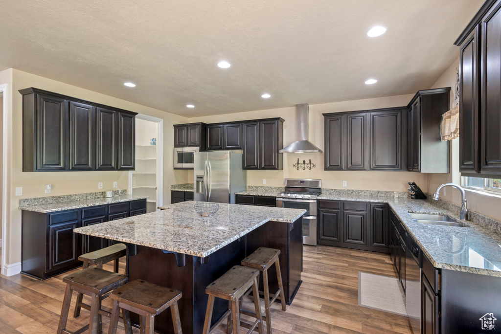 Kitchen featuring wall chimney exhaust hood, a sink, stainless steel appliances, a kitchen island, and wood finished floors